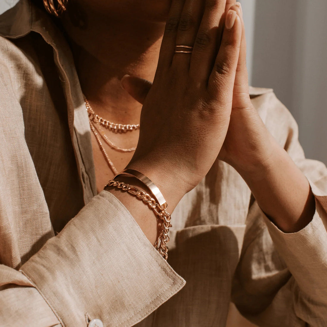 Close-up of person wearing layered gold necklaces, gold bracelets, and a gold ring on clasped hands